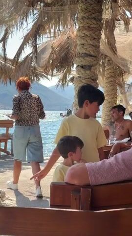 The video shows two young boys standing next to each other, both wearing yellow shirts. The older boy, on the right, looks towards something out of frame and appears to be speaking. The younger boy, on the left, is looking down and to his left. In the background, a woman in a leopard print shirt and denim shorts walks away from the camera towards a body of water. A man is visible further back, partially submerged in the water, looking towards the shore. The scene takes place outdoors, under the shade of palm trees, with a beach and mountains visible in the distance.