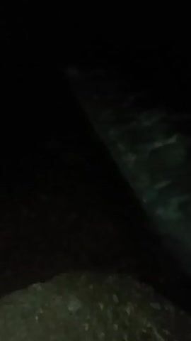 This video shows a very dark, nighttime scene next to a body of water. In the foreground, indistinct ground or foliage is barely visible. To the right and extending across the frame, a dark body of water is seen, characterized by ripples and movement, indicating a current. The camera pans slightly, focusing on the water. The overall visibility is extremely low, suggesting complete darkness apart from what little light might be reflecting off the water. The primary activity is the flow of water. There are no people or clear objects, and no visible text. The setting is an outdoor, natural environment, likely a river or stream at night in Tahlequah, United States of America.