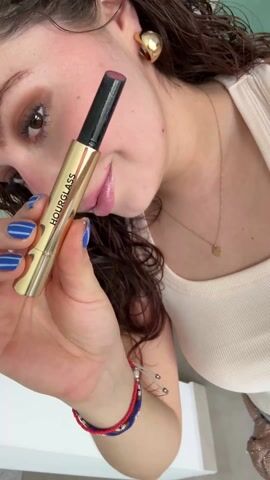 A person is shown from a high angle, holding a golden tube of Hourglass brand lip product and applying it to their lower lip. The person has fair skin, brown eyes, and curly brown hair, and is wearing gold earrings, a gold necklace, and a white tank top. They have blue nail polish on their fingernails and are wearing a red and blue bracelet on their wrist. The background is light and the floor is a light-colored tile.
