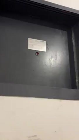 The video shows a close-up of a gray metal door or panel, possibly an access panel or service door. The door has a black frame and a white wall is visible underneath. A small, rectangular label is affixed to the center of the door with what appears to be a small, red-colored hole or light beneath it. The video appears to be taken in an indoor setting, likely a building's interior. The lighting is somewhat diffused, causing reflections on the gray painted surface of the door.