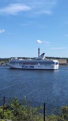 This media file captures a wide shot of the ferryboat "Silja Line" sailing on a body of water under a bright blue sky with scattered clouds. The ferry is white with blue lettering spelling out "SILJA LINE" along its side, and a distinctive blue and white logo on its funnel. The ferry is a large passenger vessel, suggesting it is likely carrying many people.

In the background, the shore is visible with buildings and a tall tower, some of which appear to be industrial or port structures. The water is a deep blue, and the overall scene suggests a clear and sunny day, likely during daylight hours. The foreground shows a wire fence and some greenery, indicating the vantage point is from land, possibly a park or viewpoint overlooking the water. The presence of the ferryboat implies travel or commerce is taking place on the waterway. The location context of Lidingö, Sweden, suggests this scene is likely in the Stockholm archipelago. There are no people clearly visible on the deck of the ferry.