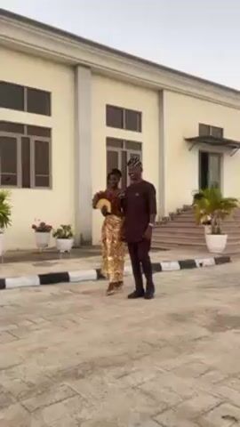 A young couple, dressed in formal Nigerian attire, walks hand-in-hand on a paved area in front of a two-story, light-colored building. The woman, on the left, wears a dark brown top with a shimmering gold skirt and gold heels, and carries a feathered fan. The man, on the right, wears a brown ensemble with a matching hat and black dress shoes. They are smiling and appear joyful as they stroll. The building behind them has evenly spaced windows and is decorated with potted plants. The pavement is marked with black and white stripes along the edge.