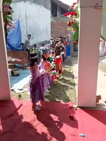 The scene shows a group of young children, mostly girls, lined up in a yard. They are dressed in various traditional costumes, including headbands and colorful garments. Each child is holding a small bowl in their hands. A few adults are visible in the background, one of whom is standing and observing the children. The surroundings include a building with brick and white walls, a blue tarp, a bench, and some cooking utensils. There are decorative flowers on a white pillar near the children. The setting appears to be an outdoor event or gathering. The ground is covered in grass and a red carpet. The overall atmosphere seems festive.