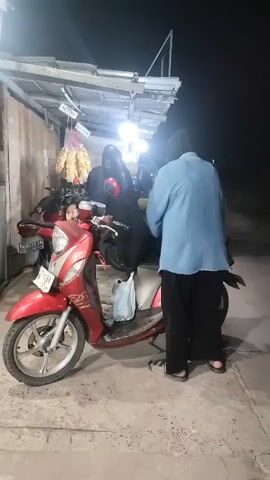 A still night scene shows a red scooter parked on a paved surface in front of a stall with a corrugated metal roof supported by metal poles. A person wearing a blue shirt and black pants stands to the right of the scooter, facing away from the camera and towards the stall, appearing to be engaged with something on the scooter. A second person, possibly a child, is partially visible behind the handlebars of the scooter, looking towards the camera. Several other motorcycles are parked behind the red scooter. A bundle of dried food items hangs from a hook on the stall. The lighting suggests artificial illumination from the stall. The ground is lit by the lights, and the background is dark. The overall atmosphere is that of a nighttime street scene at a vendor stall.