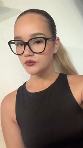 This media file features a young woman with blonde hair pulled back in a ponytail, wearing large black-rimmed glasses and a black tank top. She is posing for a selfie against a plain white background. Her makeup includes dark lipstick and mascara. Her nails are painted in bright colors, with some appearing to be a vibrant yellow and others a hot pink with glitter accents. She is looking directly at the camera with a neutral or slightly seductive expression. In some frames, she is touching her chin or cheek with her hand. There are no visible text elements in the media file. The lighting suggests it is indoors, possibly during the day or evening, with artificial light. There are no discernible cues to suggest the specific location of Beguntsi, Bulgaria, beyond the provided context. The overall mood is one of self-presentation and confidence.