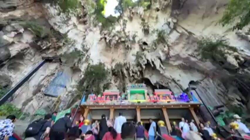 People are climbing a set of colorful stairs towards a Hindu temple structure nestled in a cave. The cave ceiling is visible, showing a large opening to the sky. There are various trees and other vegetation scattered throughout the cave. A black structure resembling a lift or hoist is visible on the left side of the stairs.  The temple structure at the top of the stairs is brightly decorated with lights and colorful ornamentation.