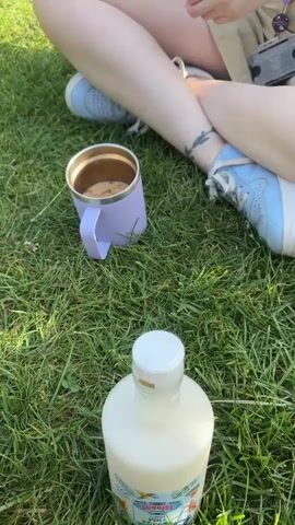 This video shows a young woman sitting on a grassy lawn in a park. She is wearing a light blue crop top, khaki shorts, and light blue sneakers. She has a lanyard around her neck with a purple strap that has "UCAS" printed on it multiple times. Her legs are crossed, and a light purple insulated mug with a beverage is to her left. A clear bottle with a white cap is in the foreground. The sun is shining, casting shadows, suggesting it is daytime, likely mid-morning or afternoon. The woman is looking off to her right, with a neutral expression. In the background, trees line the edge of the park, and other people are visible in the distance, one of whom appears to be walking. The overall scene evokes a relaxed, outdoor setting, possibly during a break or a casual outing. The presence of the UCAS lanyard might suggest she is a student attending an event or orientation related to higher education admissions.