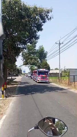 The video is a first-person perspective shot from a motorcycle, showing a street scene. In the foreground, the handlebar and a side mirror of the motorcycle are visible. The road ahead is asphalt, lined with trees and foliage on both sides. A bright red and blue jeepney, decorated with various colorful designs and lights, is parked or moving slowly on the right side of the road. Several people are visible inside the jeepney. Further down the road, other vehicles, including cars and motorcycles, are seen. The sky is clear and blue, indicating a sunny day. The overall environment appears to be a rural or semi-urban area with a mix of natural scenery and infrastructure like utility poles and wires.