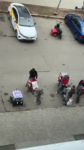 The video captures an outdoor scene with several vehicles and individuals. In the upper left, a white taxi with a yellow stripe is parked. To its right, a red scooter with a red delivery box is parked. Further right, a blue convertible is parked. In the center of the frame, four individuals on electric scooters with red delivery boxes are gathered. One individual in a black hooded jacket is standing near a black electric scooter. Three other individuals are near two other scooters. A small white delivery robot with wheels is in the lower left of the frame. The ground is paved with asphalt and some brickwork.