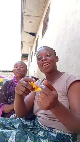 Here's a description of the video:

Two women are sitting outside, one is holding and showing a piece of yellow fruit. The woman on the right is wearing a light brown t-shirt and a blue and green patterned skirt. The woman on the left is wearing a purple top with a leopard print and a blue and green patterned skirt. They both appear to be talking or singing. The background shows a building with a white wall and visible window frames. Clothes are hanging on a line in the distance.