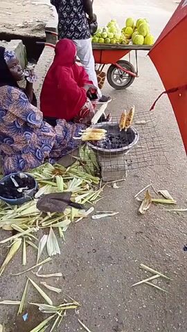 A street scene shows two women selling corn. The woman on the left is wearing a blue and purple patterned dress and a black hijab, and is drinking from a bottle. The woman on the right is wearing a red hijab and dress, and is tending to corn being grilled over a charcoal brazier. Several ears of corn are on the grill, and more are on skewers. Corn husks are scattered on the ground around them. In the background, a person in a white shirt and patterned vest stands by a cart filled with fruits, possibly melons. A red umbrella is partially visible to the right. The ground is paved with asphalt.