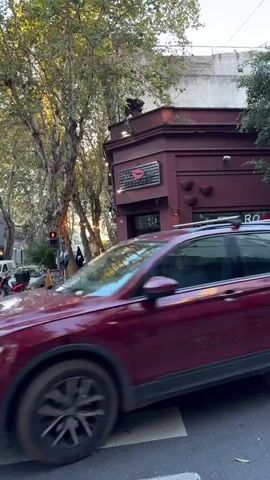 The video shows a street corner with a dark red restaurant on the corner. The restaurant is called "Ro Besa Restaurante." There are trees on either side of the street, and a crosswalk in the foreground. Cars drive by on the street. Two pedestrians are on the sidewalk, and one crosses the street. A moped is parked on the sidewalk. The sky is overcast. There is a sign outside the restaurant with the menu.