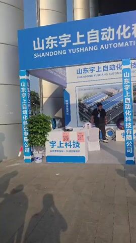 The video pans across an outdoor exhibition area with several booths. A large banner with Chinese characters and "SHANDONG YUSHANG AUTOMATION TECHNOLOGY CO., LTD." is visible at the top. In the foreground, a man in a dark shirt and pants walks past a booth displaying several colorful wheel rims. Other booths are visible in the background, with people interacting and examining displays. The environment appears to be a sunny day with shadows cast on the paved ground.