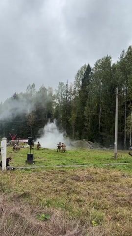 This video depicts a reenactment event set in a grassy field with a wooded area in the background. Several individuals, dressed in military-style uniforms, are positioned in the center of the frame, some holding firearms. Smoke is emanating from various points on the field, suggesting the use of pyrotechnics for dramatic effect. In the background, a red fire truck is visible, along with several informational signs. One sign, in Russian, reads "ЮРЬЕВСКИЙ РУБЕЖ" (Yuryevsky Rubezh), another reads "МЕСТО ВОИНСКОЙ СЛАВЫ РОССИИ" (Place of Military Glory of Russia), and another refers to "ИНТЕРВЕНЦИЯ (1918-1922)" (Intervention (1918-1922)). A large red star structure is also present, along with what appears to be a hedgehog anti-tank obstacle. The sky is overcast, indicating a cloudy day. The overall scene suggests a historical reenactment or a commemorative event related to military history.
