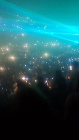 A densely packed crowd of people is illuminated by the light of cell phone screens in a concert setting. Above them, colorful stage lights in shades of blue, green, and white sweep across the space, creating a dynamic atmosphere. The focus is on the crowd's interaction with the performance, evident through the raised phones capturing the event. The environment is dark, emphasizing the contrast between the light sources and the shadows cast by the audience.