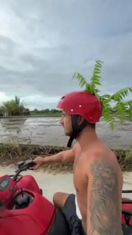 The video shows a shirtless man riding a red ATV. He is wearing a red helmet adorned with green fern-like leaves. The man has a beard and a tattoo on his right arm. The ATV is moving on a narrow concrete path beside flooded rice paddies under a cloudy sky.