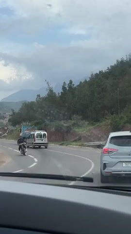 The video captures a first-person perspective from inside a vehicle driving on a road in Taljarft, Morocco, under an overcast sky. The road winds through a mountainous and hilly landscape, with green vegetation and trees covering the slopes.

Several vehicles are ahead on the road. Initially, a motorcyclist is visible on the left side of the road, followed by a white van and then a truck with a blue bed. Directly in front of the recording vehicle, a silver Renault Clio is driving in the right lane. The Clio's Moroccan license plate, "78771 | 26", is clearly visible, as is the "CLIO" emblem on its rear.

On the left side of the road, a building with a partially visible sign reading "ATELIER DU POTIE" (Potter's Workshop) is nestled into the hillside. The scene depicts a drive through a rural, mountainous region.