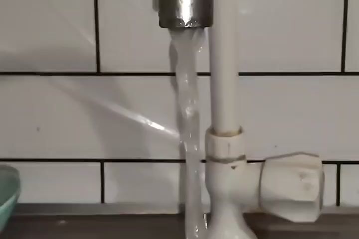 The video shows water flowing from a faucet into a sink. The faucet is silver and the pipe leading to it is white. The background is a white tiled wall with black grout. The sink appears to be made of a dark material, possibly stainless steel or granite. The water stream is somewhat broken, indicating it might be aerated or the flow is not entirely smooth. There is a light blue object visible to the left of the faucet, partially obscured by the water. The overall scene suggests a kitchen or bathroom sink.