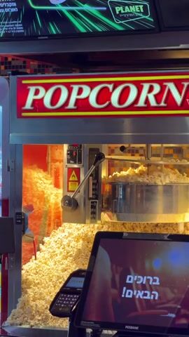 A popcorn machine is shown from a medium close-up. The machine has a red illuminated sign that says POPCORN in white letters with a yellow outline. The front of the machine is open, revealing a large pile of freshly popped popcorn. Some popcorn is still falling from the top into the pile. There are controls on the front of the machine. In the foreground, a dark tablet device with a payment terminal is visible. The screen of the tablet displays text in Hebrew that translates to "Welcome the guests!". The background shows a blurred image of what appears to be a movie theater ceiling with green lighting. The overall environment suggests a concession stand at a movie theater or entertainment venue.