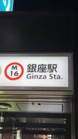 The video begins with a close-up of a sign for the Ginza Station (Ginza Sta.), marked with "M6" and a Japanese inscription. The camera then tilts upwards to show a large building with ornate decorations lit up at night. The building has a large illuminated archway with intricate designs, as well as advertisements and the logo of Mitsukoshi.