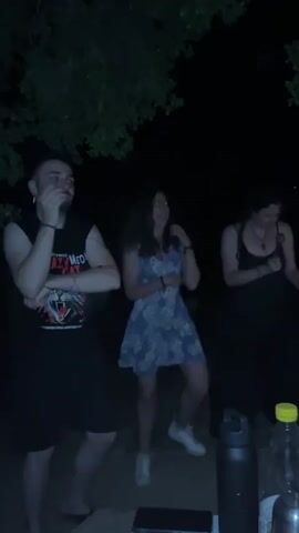 Three people are dancing outdoors at night. The person on the left is a man with a beard wearing a black tank top, black shorts, and a cigarette in their mouth. They dance with their right arm raised and left arm bent. The person in the middle is a woman with long dark hair wearing a patterned blue and white dress and white sneakers. She dances with both arms raised. The person on the right is a woman with long dark hair wearing a black tank top and a long black skirt. She is holding an object in her right hand and swaying. A dark water bottle with a yellow cap and a black water bottle are on a surface in the foreground. Trees are visible in the background.