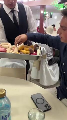 The video shows two waiters circulating through a reception hall offering guests various popsicles. The first waiter wears a black vest, white shirt, and tie. He is holding a silver tray that contains popsicles of various colors and flavors. A guest with a cowboy hat takes one of the popsicles. The camera follows the waiter as he continues to serve the guests. The video then cuts to a second waiter wearing khaki pants and a blue and white plaid shirt, holding another tray of popsicles. Another guest with a white apron takes a popsicle from the tray. Several tables are visible in the background, along with other guests and staff members. The reception hall appears well-lit, with decorations and ambient noise.