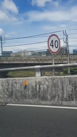A video shows a view from a vehicle traveling along a highway. The camera is focused on the right side of the vehicle, offering a perspective of the scenery passing by. The foreground is dominated by a concrete barrier with a white metal railing on top, featuring occasional orange markers. Beyond the barrier, a grassy area and vegetation can be seen. In the middle ground, an elevated highway or bridge is visible, supported by concrete pillars. This structure appears to be a transportation infrastructure, possibly for trains or vehicles, as indicated by the presence of overhead wires and utility poles. In the background, several multi-story buildings are sparsely scattered, suggesting an urban or suburban environment. The sky is partly cloudy with patches of blue. The video captures the linear movement of the vehicle as it progresses along the road.