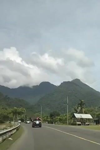 The video shows a view from a vehicle driving on a road with lush green mountains in the background. Several cars and motorcycles are visible on the road. The sky is partly cloudy, with large white clouds. There are trees and vegetation on both sides of the road, as well as some buildings and utility poles. The overall scene suggests a scenic drive through a rural or mountainous area.
