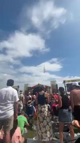 Here is a description of the video:

The video shows a crowd of people gathered in an outdoor setting, likely a music festival or concert. In the background, there's a stage set up with screens. A flag is flying in the sky above the crowd.  People are dressed in casual summer attire and some are wearing hats, seemingly enjoying the event. The sky is bright blue with scattered clouds.