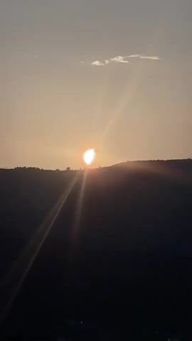 The video shows a time-lapse of a sunset over a silhouetted landscape. The sun is low in the sky, casting long rays of light. The sky is a pale orange and pink, with a few clouds visible. The landscape below is dark and indistinct. The sun appears to be setting behind a hill or mountain range. There are no people or objects clearly visible in the foreground or midground. The overall impression is one of a peaceful and serene natural scene.