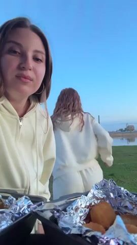 Here's a concise description of the video:

A young woman is eating outside near a body of water, seemingly at a picnic. She's wearing a light-colored hooded sweatshirt and hoop earrings. A tray in front of her holds nuggets wrapped in aluminum foil. She dips one into a small container of sauce and eats it, looking at the camera. In the background, another person in a matching outfit walks near the water. The sky is blue.
