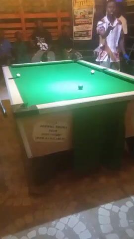 The video shows a pool table in what appears to be a bar or recreational area. Several people are visible in the background, seated and standing. The pool table has a green felt surface with several balls on it, including the cue ball and a black ball. The table has a white border and a green cloth hanging from its sides. A sign is attached to the side of the table with some text and a phone number. The lighting suggests an indoor setting, possibly in the evening. The camera is positioned at a low angle, looking up at the pool table.