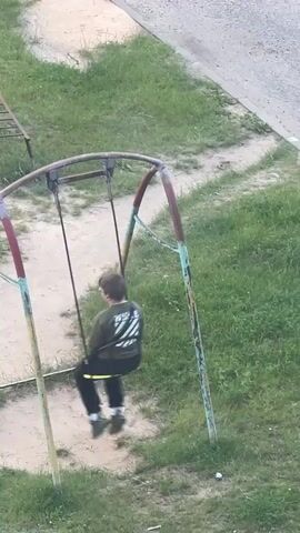 The video shows an overhead view of a playground scene in Asipovichy, Belarus. The main subjects are two young boys. One boy is actively swinging on a worn, metal swing set, its frame showing signs of rust and faded paint in red, green, and yellow. He is wearing a dark green long-sleeved shirt with a graphic design on the back that appears to have white stripes and possibly text, black pants, and grey sneakers. His head is down, and his movement suggests he's enjoying the swing.

The second boy is visible in the background, walking away from the swing set. He is wearing a green t-shirt, dark shorts, and dark sneakers. The ground around the swing set is a dirt path, worn smooth by use, bordered by patches of green grass. The setting appears to be an outdoor public space, likely a residential area given the presence of a paved surface and the overall layout.

The lighting suggests it might be late afternoon or early evening, with soft, diffused light. There are no strong shadows, and the overall atmosphere seems calm and ordinary, depicting a typical moment of childhood play. The focus remains on the children's activity and the simple playground equipment.