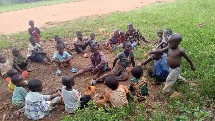 A group of about 20 children are sitting in a circle on the ground in a grassy area. Some are smiling and looking towards the center of the circle, while others are looking around. The children are dressed in various clothing styles. A woman is standing near the edge of the circle with a baby strapped to her back. A dirt road is visible in the background.