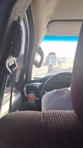 The video shows an interior view from the back seat of a parked car. The driver, wearing a white shirt, is visible behind the steering wheel. An orange can with writing on it sits in the compartment in front of the driver. Through the windshield, a large truck and other vehicles are visible on a dusty road. The sun is bright, creating a glare on the windshield. The interior of the car includes a brown cloth seat, seatbelt, and other standard car features. Loud music is audible.