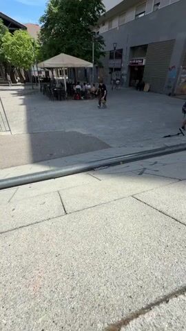 A young male skateboarder wearing a black t-shirt and brown pants grinds on a metal rail in a paved outdoor area. The skateboarder is in motion and appears to be mid-trick. In the background, there are trees, buildings, tables, and chairs, suggesting an urban or public space. The lighting indicates it is daytime with clear skies.