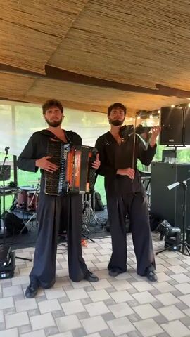 TWO YOUNG MEN ARE PERFORMING MUSIC OUTDOORS UNDER A COVERED PATIO. THE MAN ON THE LEFT IS PLAYING AN ACCORDION AND THE MAN ON THE RIGHT IS PLAYING A VIOLIN. BOTH MEN ARE WEARING BLACK SHIRTS AND WIDE-LEGGED BLACK PANTS. THE MAN ON THE LEFT IS WEARING A SHIRT WITH A HOODIE AND THE MAN ON THE RIGHT HAS A BOW IN HIS HAND WHILE PLAYING THE VIOLIN. MUSIC EQUIPMENT SUCH AS SPEAKERS AND DRUMS ARE VISIBLE IN THE BACKGROUND.