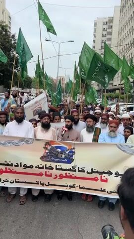 This is a vertical, medium shot video of a protest taking place in a city. A large group of men, predominantly wearing traditional Islamic attire such as turbans and long beards, are gathered on a street. Many of them are holding green flags with white Arabic script that appears to say "SUC" and potentially "Pakistan" or "Federation."

In the foreground, a large banner is stretched across the width of the frame, held by several men. The banner has images of chains and a padlock over a TV logo that reads "GNN" and "GEO." Below this, there is text in Urdu script, which translates to a protest against the Geo and GNN channels. One man in the center, wearing a turban, is speaking into a microphone, likely addressing the crowd.

The backdrop of the protest includes modern, tall buildings, suggesting an urban setting. The weather appears to be overcast, and the time of day is likely daytime. The overall mood seems to be one of protest and strong sentiment, with the participants appearing serious and united in their cause. The visual elements, including the flags, banner, and the man speaking, all point to a public demonstration against media outlets. The location is identified as Karachi Division, Pakistan.