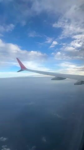 The video shows a view from an airplane window, looking down at clouds and the earth below. The airplane wing is visible in the foreground, with a red tip on the end. The sky is blue with scattered white clouds. The earth below is covered in a thick layer of clouds, obscuring most details, but some faint land features and bodies of water can be discerned. The lighting suggests it is daytime, possibly sunrise or sunset due to the soft light. The video is short and shows no people or direct actions, focusing entirely on the aerial landscape.