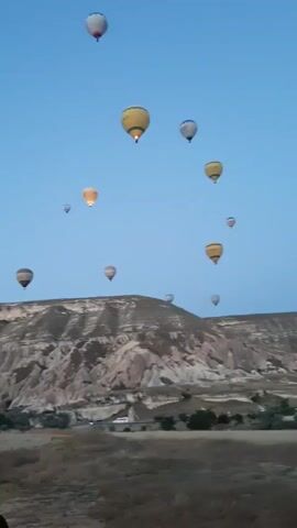 This is a landscape video taken from the ground, showing a clear, blue sky with multiple hot air balloons ascending and floating in the distance. The balloons are of various colors, including yellow, white, and orange, with some illuminated from within, suggesting it is either dawn or dusk. Below the sky, there is a rugged, mountainous terrain with a dry, grassy field in the foreground. A white van is parked on the left side of the frame, and the back of a person's head and shoulder are visible in the lower left corner, indicating the perspective of someone filming the scene. The overall atmosphere is serene and picturesque, capturing a common tourist activity in a scenic location.