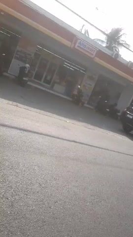 The video pans across a street scene. In the foreground, the asphalt road is visible with a long shadow cast across it. In the background, there are buildings, including what appears to be a convenience store with an "Alfamart" sign. Several motorcycles are parked outside the store. Further down the street, there are houses and more shops. The sky is visible, suggesting it is daytime. The camera appears to be mounted on a moving vehicle, possibly a motorcycle, as indicated by the reflection of a mirror in the bottom right corner of the frame in some segments. The overall impression is a typical street view in a residential or commercial area.