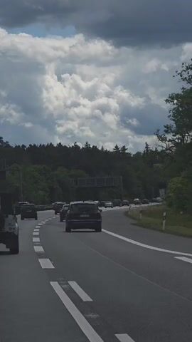 The video shows a highway scene, with several cars driving on a multi-lane road. The road is lined with white markings, and the traffic appears moderate.  Trees are visible lining the side of the road. The sky is dominated by large, fluffy, grey and white clouds. The clouds fill a significant portion of the frame, suggesting potentially unstable weather conditions. A jeep is visible in the left lane.
