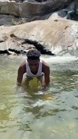 A young man is submerged in a body of water at the base of a rocky waterfall. He wears a white tank top and is waist-deep in the greenish water. Initially, he is kneeling, then he stands up, his arms extended, before turning and swimming a few strokes. The waterfall cascades down gray rocks in the background.