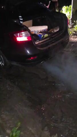 A black car is stuck in mud at night. Smoke is coming from the exhaust pipe. The trunk of the car is open, and various items are visible inside. The ground is covered in thick mud and puddles. The scene appears to be in a rural area with tall grass and a dark sky. The car's taillights are illuminated, indicating it is running. The overall impression is one of a vehicle struggling to move in difficult terrain.