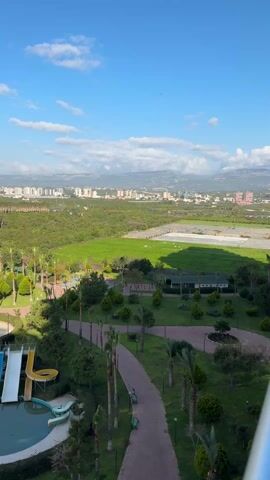 The video shows a high-angle view of a park or resort area, with a partial view of a city in the distance under a blue, partly cloudy sky. In the foreground, there is a small water park area with two water slides emptying into a pool. The park features a network of paved walking paths lined with palm trees, bushes, and green grass. Beyond the park is a large, flat grassy area with some trees. The city in the background appears to be densely populated with buildings of various heights. Mountains are visible in the far distance, beneath the skyline.