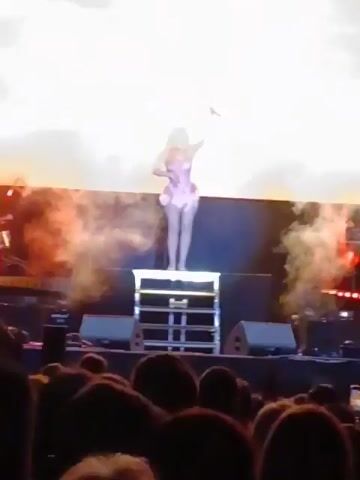A woman is standing on a small elevated, lit platform, wearing a light pink outfit. The background is a large screen with light flares. She seems to be at a performance or show, as there are other lights and possibly smoke surrounding the stage. The camera is angled up at the woman.