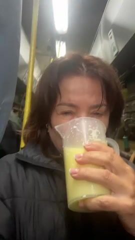 A woman with shoulder-length brown hair, wearing a black puffer jacket, is in the foreground, holding a clear plastic pitcher filled with a pale yellow liquid to her mouth. Some of the liquid is spilling down her chin and onto her jacket. Her eyes are wide and she is making a facial expression that could be interpreted as surprise or amusement. In the background, out of focus, is another person with blonde hair wearing glasses and a black jacket. The environment appears to be indoors, possibly a bus or a train, with fluorescent lighting and poles visible. There is also what appears to be some sort of equipment or machinery in the background. The video is approximately four seconds long.