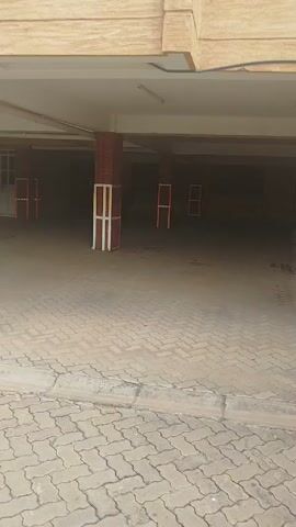 The video shows a parking garage under a building. A white SUV is parked in one of the spaces. There are potted plants near the supporting columns. The ground is paved with interlocking bricks. The camera pans up to show the building with white-framed windows and a closed black gate. The sky is overcast.