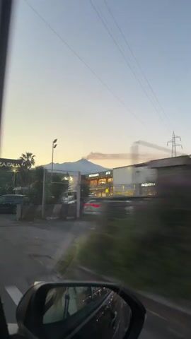 The video shows a car traveling on a road near Piazza Italia and a McDonald's. In the distance, a snow-capped mountain with smoke rising from its peak is visible. Palm trees and other foliage are present along the roadside. The sky is a clear, pale blue. There are parked cars and other vehicles moving on the road. The view is from inside the car, capturing the roadside through the window and the side mirror.