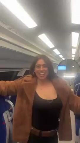 A young woman stands in the aisle of a train and dances. She has brown hair, and is wearing a brown jacket, black top, and shorts with tights. She is wearing large hoop earrings and a belt. She is smiling and sticking her tongue out. The train is empty except for her. The seats are blue and the walls are white. The ceiling has bright fluorescent lights.
